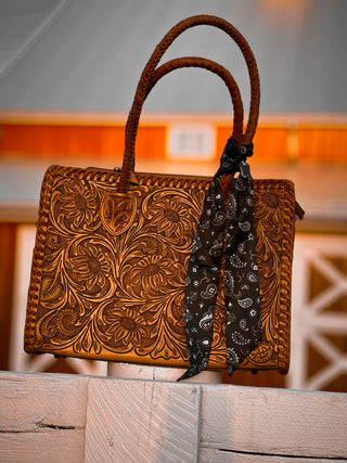Brown leather handbag with floral patterns and a bandana on a wooden surface.