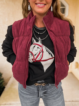Person wearing a red corduroy vest over a black shirt with a horse graphic, standing indoors.