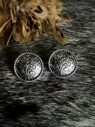 Pair of silver circular earrings with intricate western designs on a textured surface.