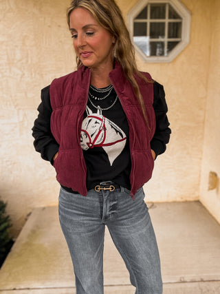 Woman wearing a black jacket with a horse design, maroon vest, and gray jeans outdoors.
